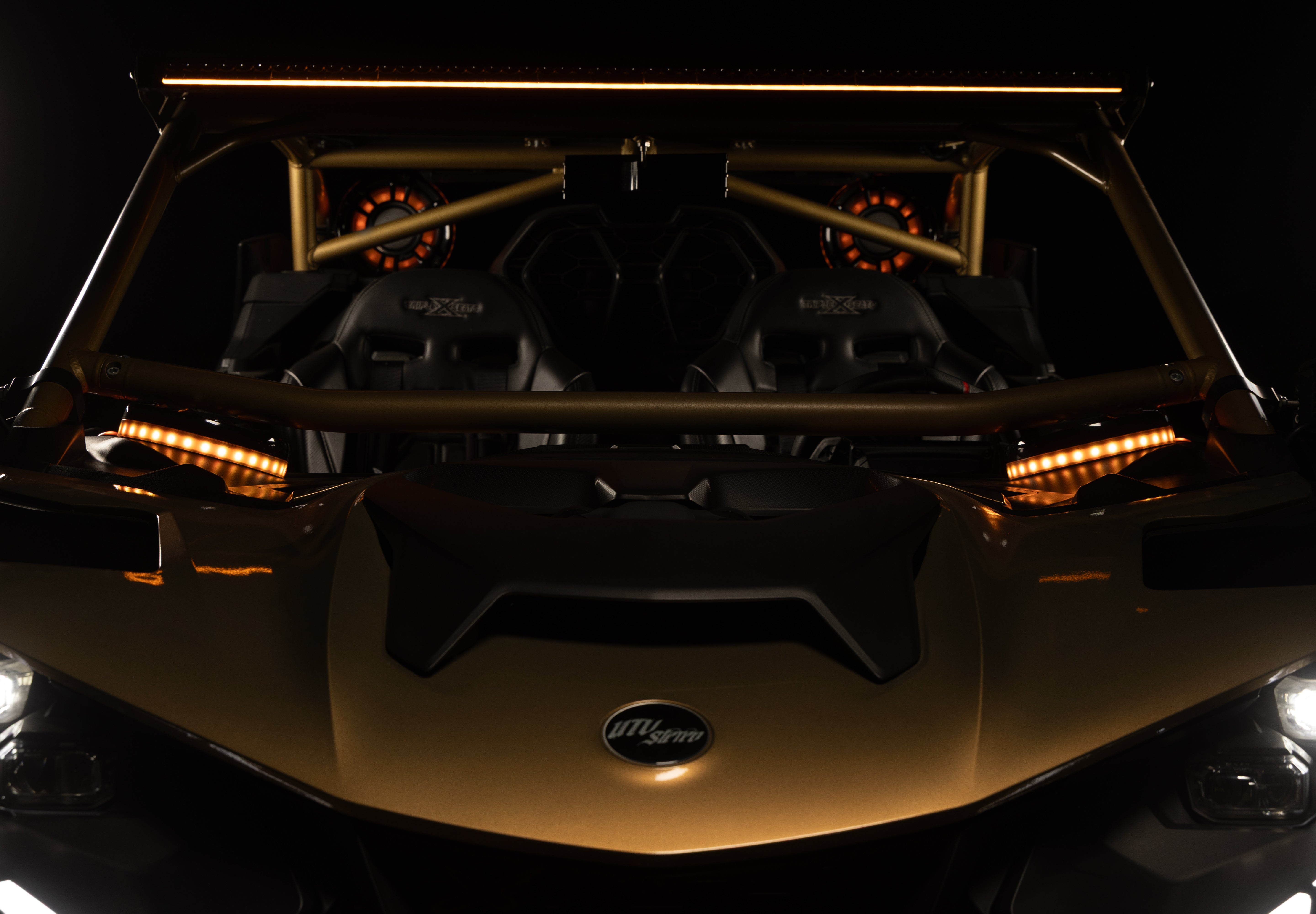 Front view of a gold and black UTV with a roll cage, dual racing seats, and amber LED light bars on a dark background.