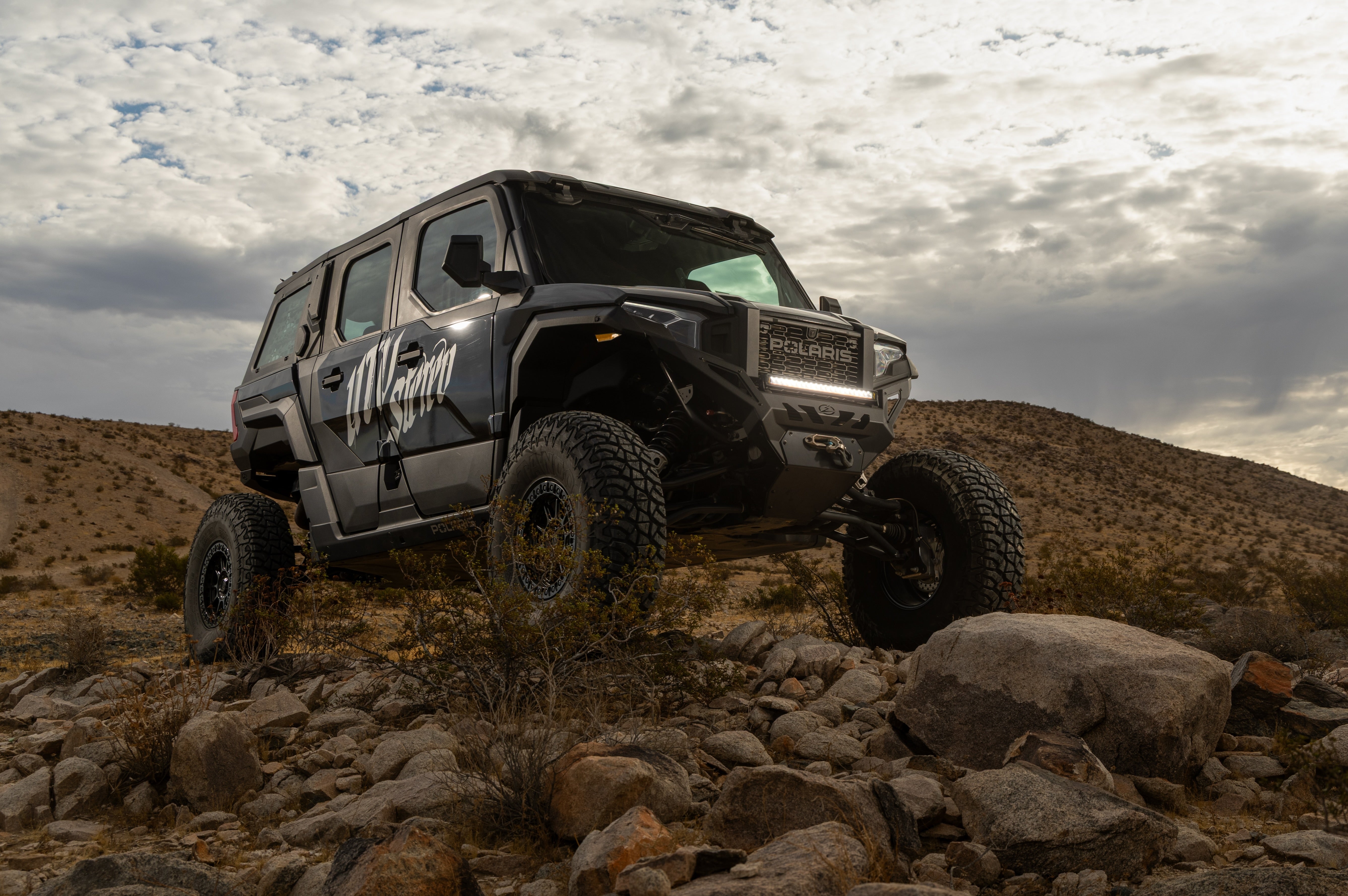 A black off-road Polaris Xpedition with large knobby tires and custom graphics, climbing a rocky desert hill under a cloudy sky.