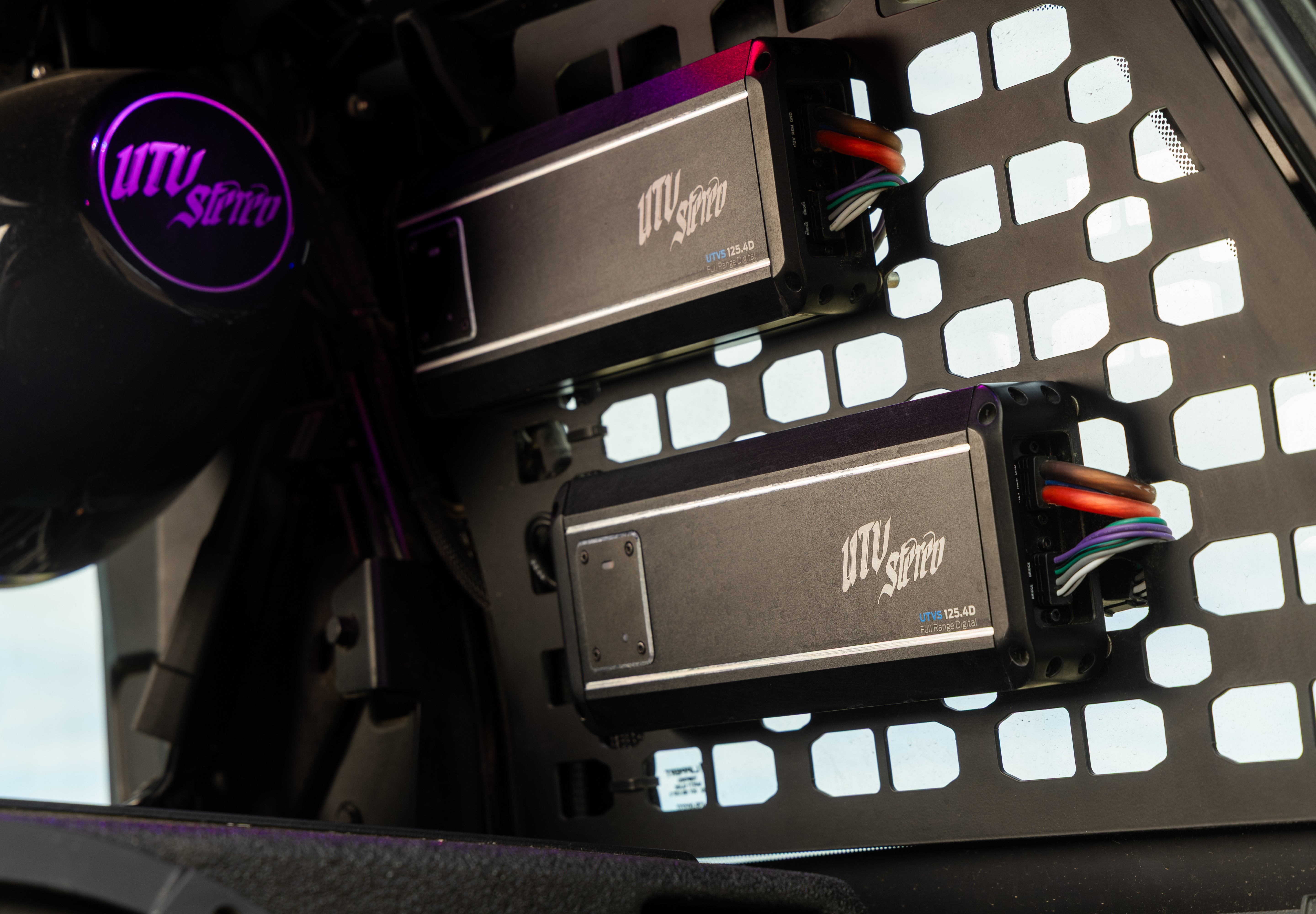 Two black UTVS electronic controller units mounted on a black metal frame with square cutouts, featuring multicolored wiring and a purple UTVS logo visible on a nearby circular component.