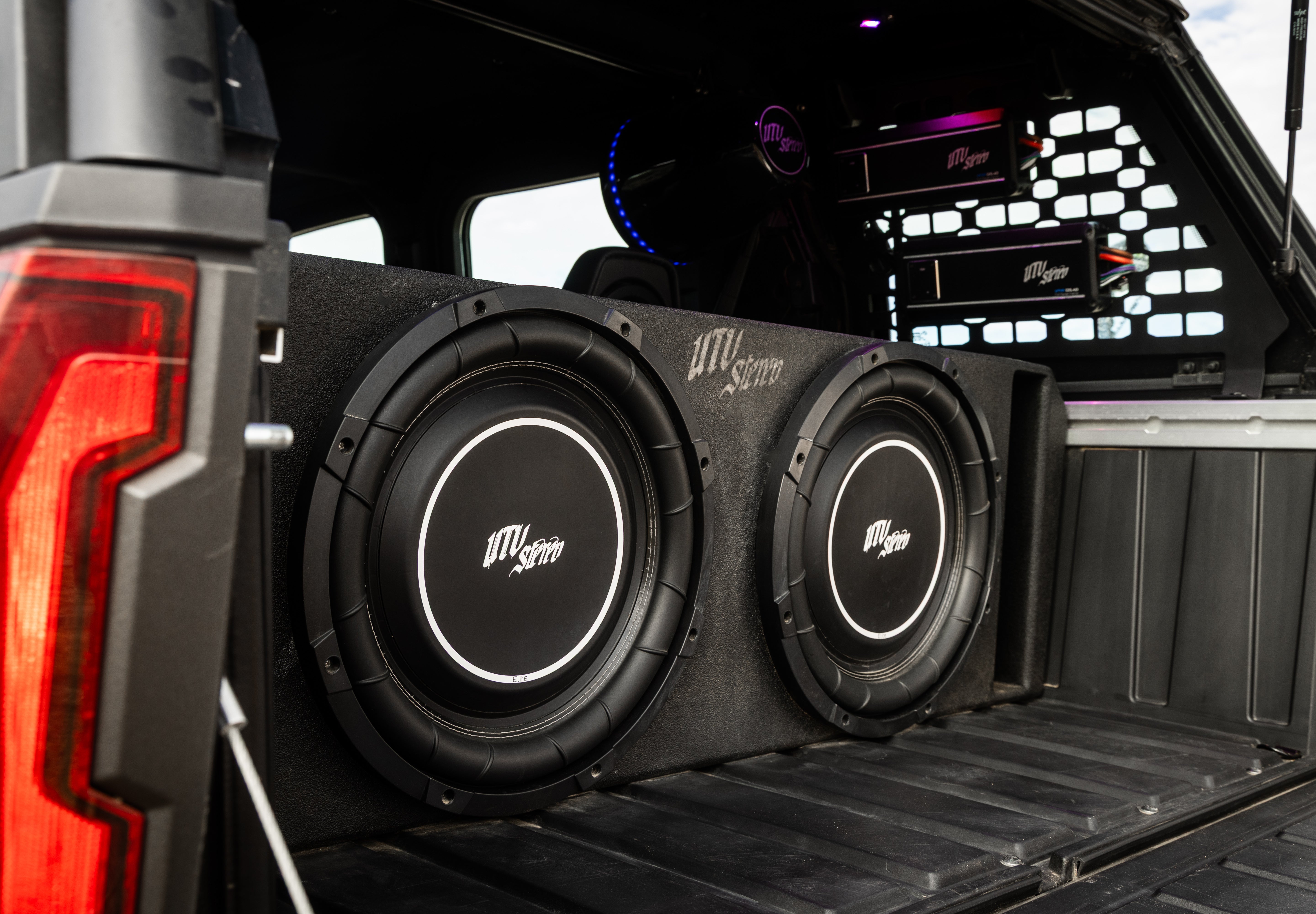 A pair of large black UTVS car subwoofers installed in a custom black speaker box inside the bed of a pickup truck.