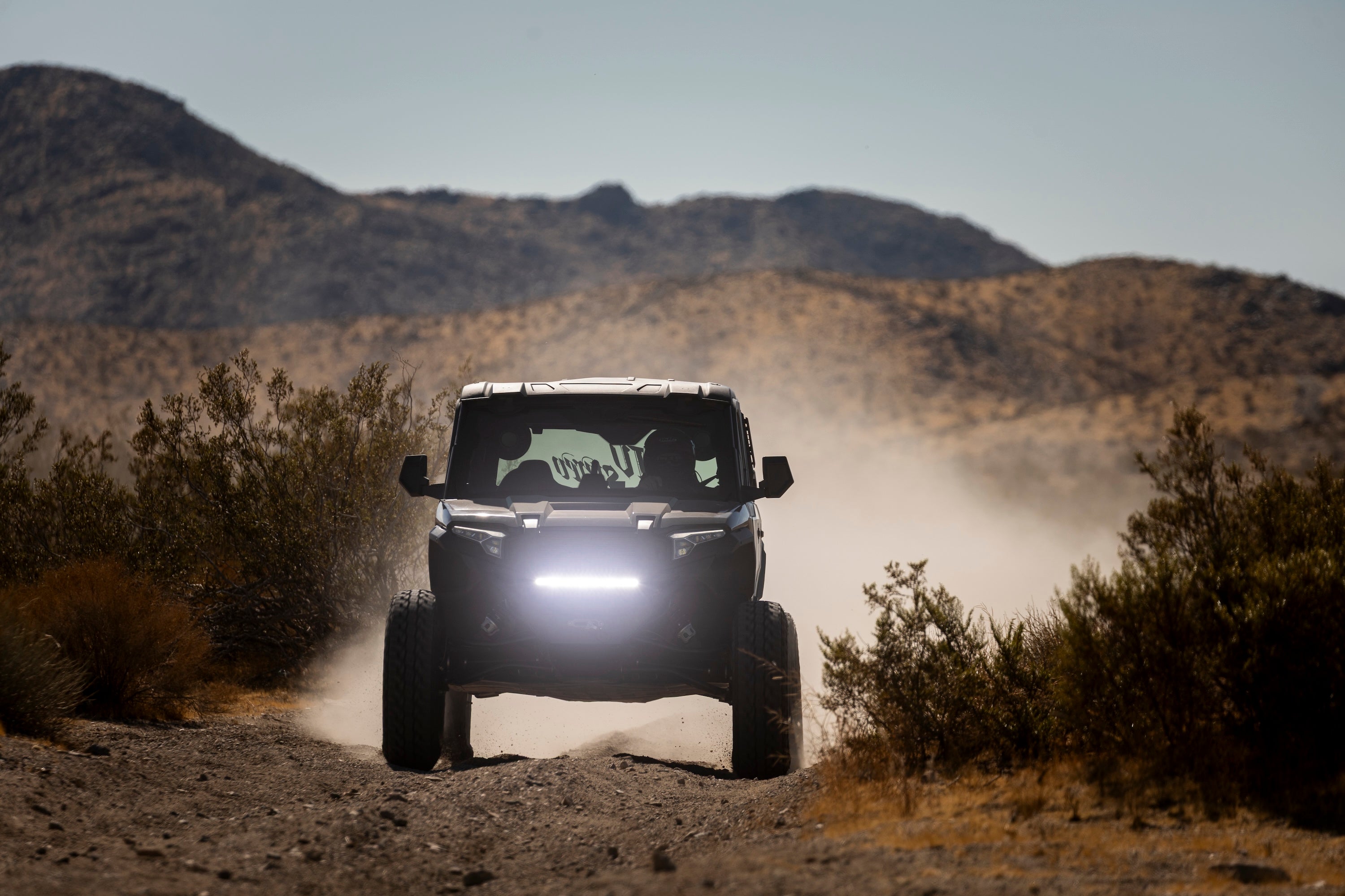 A black off-road vehicle driving toward the camera on a dusty dirt trail with mountains in the background.