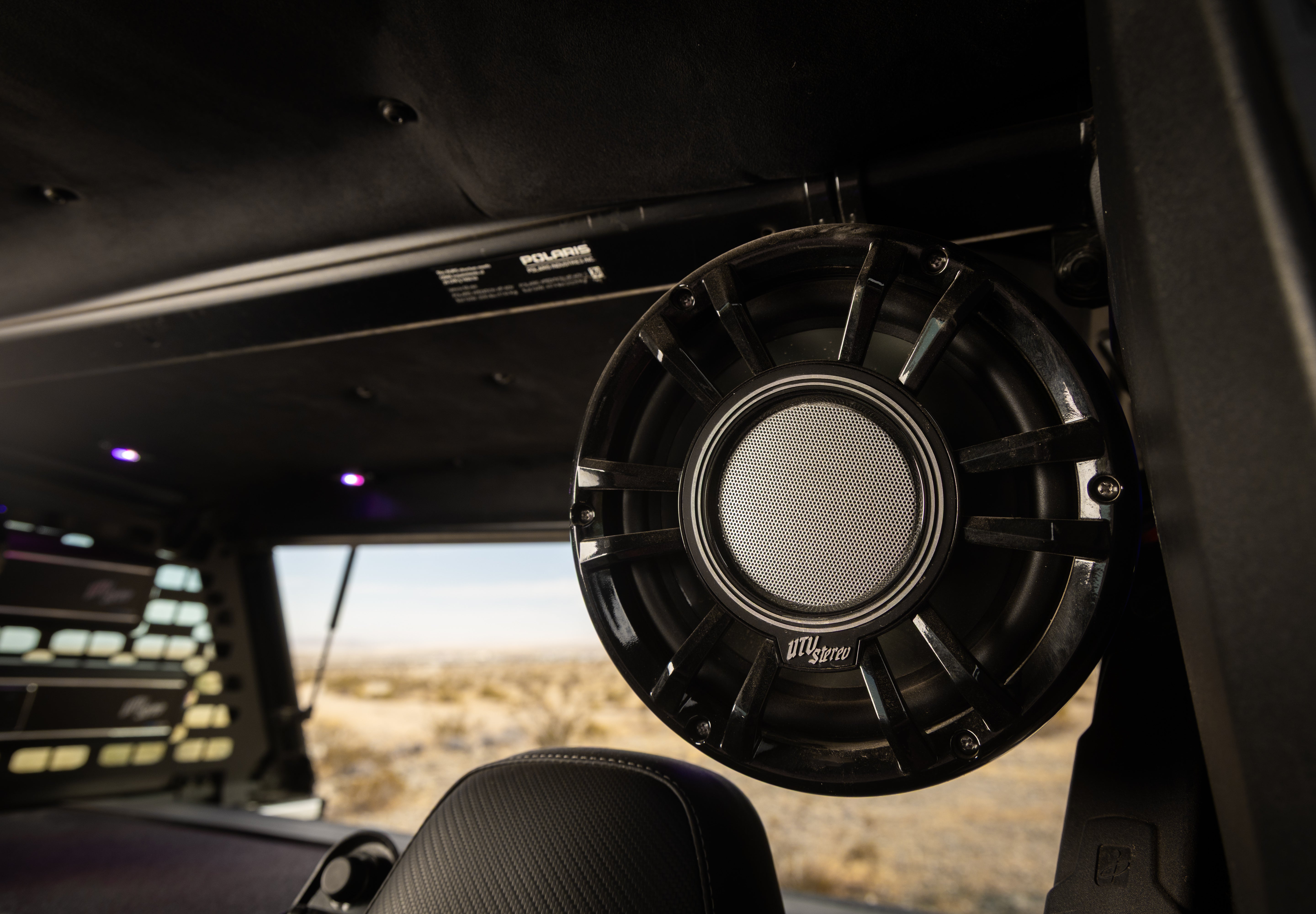 A close-up of a black UTVS speaker mounted inside a vehicle cabin, with the desert landscape visible through the windshield.